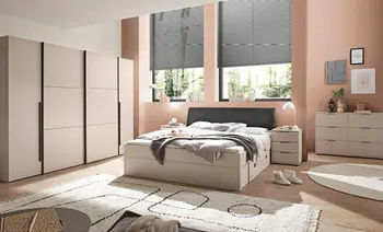 Schlafzimmer mit hellem Stauraumbett und grauem Kopfteil, links breiter Kleiderschrank, rechts Kommode und Nachttische vor apricotfarbenen Wänden und Fenstern.